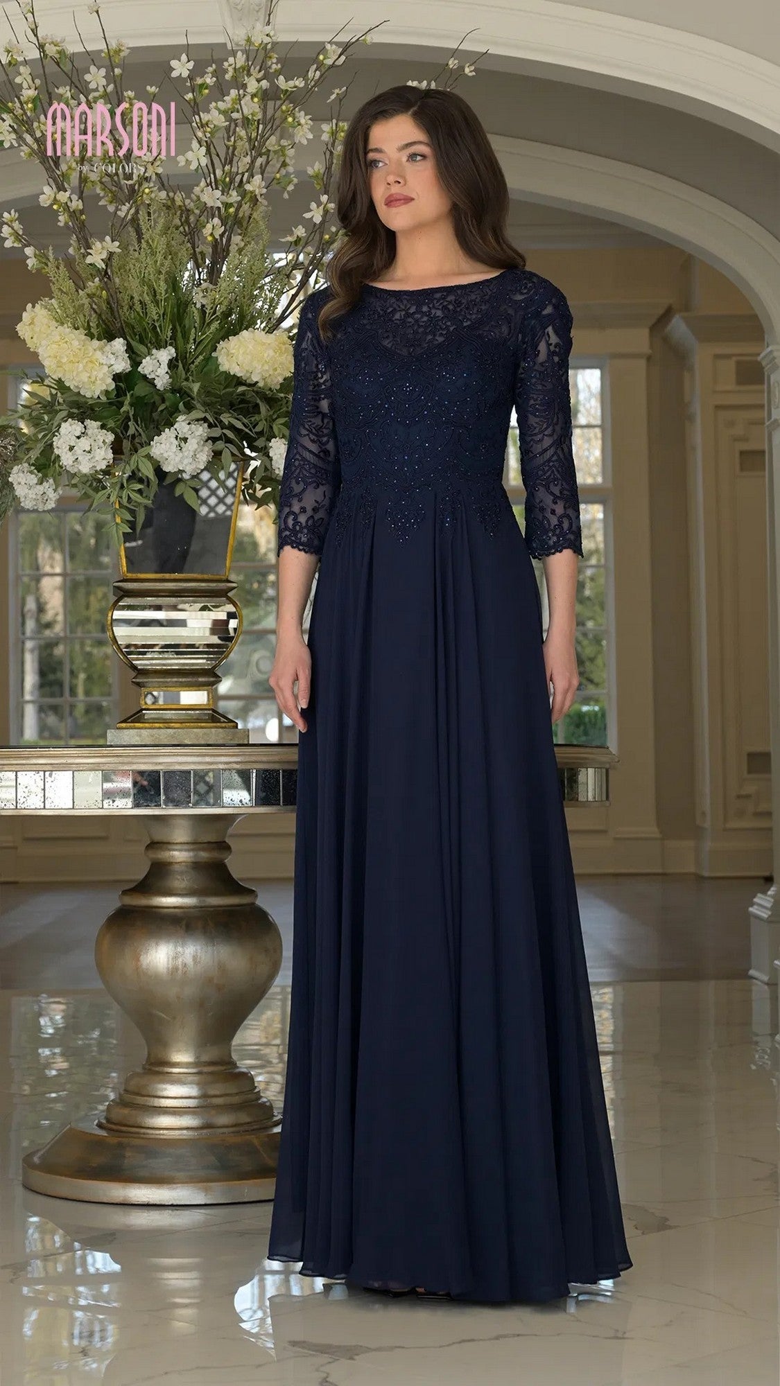 A woman wearing the navy Marsoni M238SL long formal dress, featuring a chiffon A-line skirt and beaded bodice, stands indoors beside a large floral arrangement and gold pedestal table.